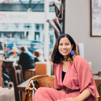 Eine Frau mit langen, dunklen Haaren sitzt in einem Café, trägt ein rosafarbenes Outfit und große Ohrringe. Im Hintergrund sind verschwommene Silhouetten von Personen und Möbeln zu sehen.