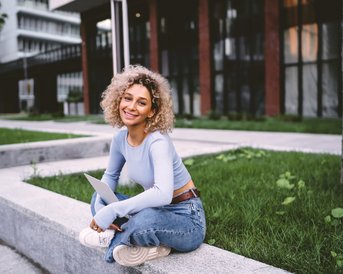 Junge Frau mit lockigem Haar sitzt auf einer Steinbank, trägt ein hellblaues Oberteil und Jeans. Sie hält ein Laptop und lächelt, während sie in einem modernen, urbanen Umfeld posiert.