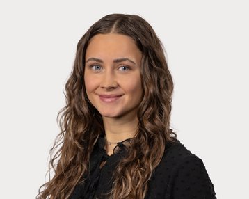 Eine junge Frau mit lockigem, braunem Haar und blauen Augen trägt ein schwarzes Oberteil und lächelt freundlich in die Kamera.