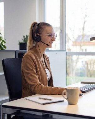 Eine Frau mit Headset sitzt an einem Schreibtisch und arbeitet am Computer, während sie in einem modernen Büro umgeben von Pflanzen ist.