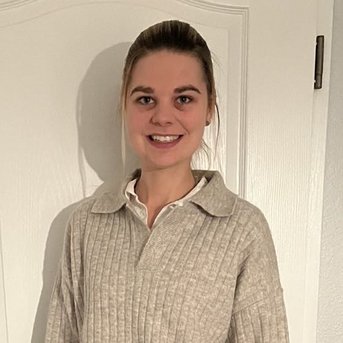 Eine Person steht vor einer weißen Tür, trägt einen beigen, gestreiften Pullover und schwarze Lederhosen. Sie hat kurze, blonde Haare und lächelt freundlich.