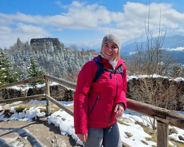 Eine Person in roter Jacke steht an einem Holzgeländer, umgeben von schneebedeckten Bäumen und einer Berglandschaft. Im Hintergrund ist eine Burgruine sichtbar.