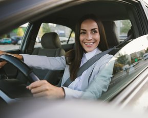 Eine Frau mit langen Haaren sitzt am Steuer eines Autos und lächelt, während sie in eine Richtung schaut. Sie trägt einen Anzug und hat den Sicherheitsgurt angelegt.