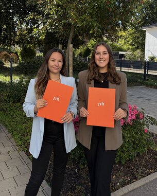 Zwei Frauen stehen im Freien und halten orangefarbene Ordner mit der Aufschrift "srh" in den Händen. Im Hintergrund sind Bäume und ein Gebäude sichtbar.