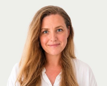 Eine Frau mit langen, lockigen Haaren und einem freundlichen Gesichtsausdruck trägt ein weißes Hemd. Der Hintergrund ist neutral.