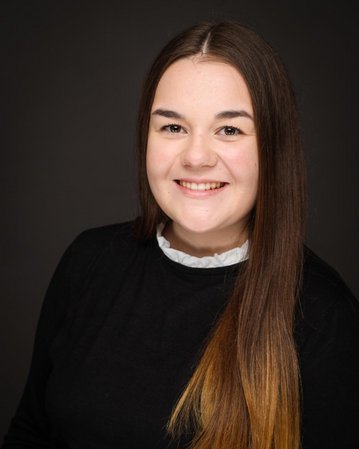 Junge Frau mit langen, glatten Haaren und einem freundlichen Lächeln, trägt ein schwarzes Oberteil mit einem weißen Kragen.