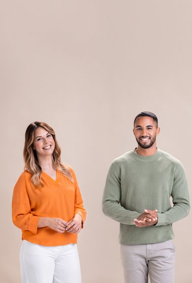 Zwei Personen stehen nebeneinander, eine Frau in einem orangefarbenen Pullover und ein Mann in einem grünen Pullover. Beide lächeln und wirken freundlich, vor einem neutralen, hellen Hintergrund.