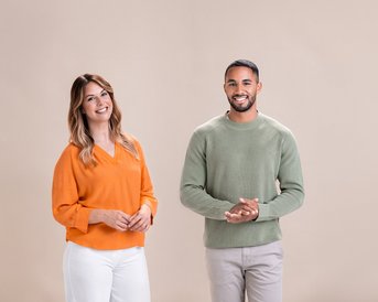 Zwei Personen stehen nebeneinander, eine Frau in einem orangefarbenen Pullover und ein Mann in einem grünen Pullover. Beide lächeln und wirken freundlich, vor einem neutralen, hellen Hintergrund.