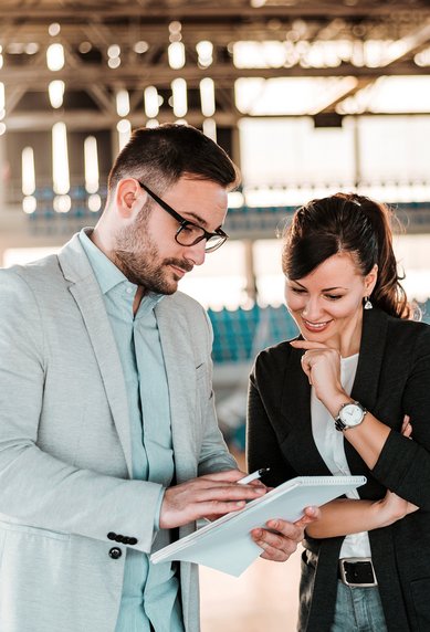 BWL und Sportmanagement Bachelor Ein Mann und eine Frau stehen in einem Sportstadion und betrachten gemeinsam ein Tablet. Beide scheinen in ein Gespräch vertieft zu sein, während im Hintergrund leere Sitzplätze sichtbar sind.