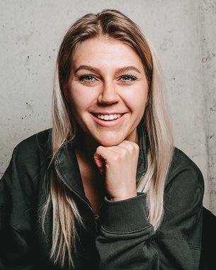 Eine lächelnde Frau mit langen, blonden Haaren sitzt in einem schwarzen Stuhl und stützt ihr Kinn auf die Hand. Der Hintergrund besteht aus einer grauen Wand.