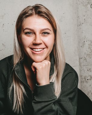 Eine lächelnde Frau mit langen, blonden Haaren sitzt in einem schwarzen Stuhl und stützt ihr Kinn auf die Hand. Der Hintergrund besteht aus einer grauen Wand.