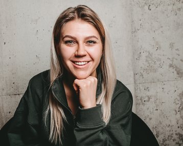 Eine lächelnde Frau mit langen, blonden Haaren sitzt in einem schwarzen Stuhl und stützt ihr Kinn auf die Hand. Der Hintergrund besteht aus einer grauen Wand.