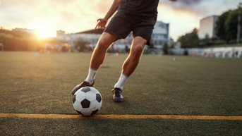 Ein Fußballspieler tritt einen Ball auf einem Rasenplatz bei Sonnenuntergang. Die Szene zeigt den Spieler in Bewegung, während er sich auf das Spiel konzentriert.