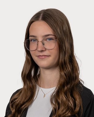 Eine Person mit langen, lockigen Haaren und Brille, die in einem neutralen Hintergrund steht. Sie trägt ein schwarzes Oberteil und hat einen neutralen Gesichtsausdruck.