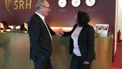 Zwei Personen in formeller Kleidung stehen an einem Empfangstresen und unterhalten sich. Im Hintergrund sind Uhren und Mitarbeiterinnen sichtbar. Das Logo „SRH“ ist an der Wand angebracht.