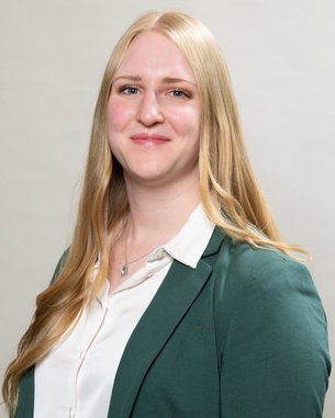 Eine blonde Frau mit langen Haaren trägt ein grünes Blazer und ein weißes Hemd. Sie steht vor einem neutralen Hintergrund und lächelt leicht.