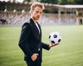 Ein Mann in einem schwarzen Anzug hält einen Fußball in der Hand, während er auf einem Sportfeld steht.