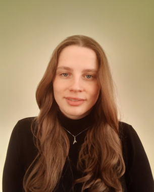 Eine junge Frau mit langen, lockigen Haaren trägt einen schwarzen Pullover und eine Halskette. Sie schaut direkt in die Kamera vor einem sanften, einfarbigen Hintergrund.