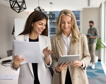 Zwei Geschäftspersonen in Anzügen betrachten gemeinsam ein Tablet in einem modernen Büro. Ein Tisch mit Laptops und Stühlen ist im Hintergrund sichtbar.
