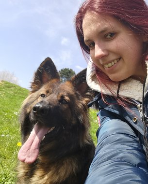 Eine Person mit rotem Haar lächelt in die Kamera, während ein Hund mit langem, braunem Fell neben ihr sitzt und den Mund offen hat. Beide sind im Freien auf einer grünen Wiese.