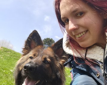 Eine Person mit rotem Haar lächelt in die Kamera, während ein Hund mit langem, braunem Fell neben ihr sitzt und den Mund offen hat. Beide sind im Freien auf einer grünen Wiese.