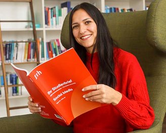 Eine Frau mit langen, dunklen Haaren sitzt in einem grünen Sessel und hält ein rotes Buch mit dem Titel "Ihr Studienführer Social Media". Sie lächelt und trägt einen roten Pullover.