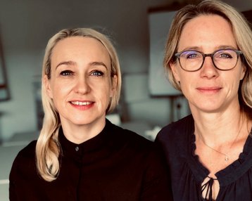 Zwei Frauen sitzen nebeneinander, eine mit blonden Haaren und die andere mit hellen, langen Haaren. Beide tragen formelle Kleidung und schauen direkt in die Kamera.