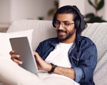 Mann mit Kopfhörern sitzt auf einer Couch und nutzt ein Tablet, während er entspannt lächelt.