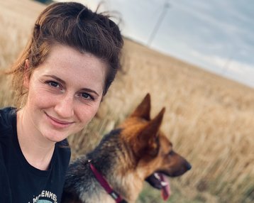 Eine Person mit lockigem Haar lächelt in die Kamera, während ein Hund neben ihr sitzt. Im Hintergrund erstreckt sich ein Feld mit hohem Gras.