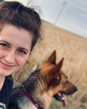 Eine Person mit lockigem Haar lächelt in die Kamera, während ein Hund neben ihr sitzt. Im Hintergrund erstreckt sich ein Feld mit hohem Gras.