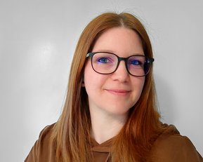 Eine Person mit langen, braunen Haaren und Brille lächelt in die Kamera, trägt einen braunen Hoodie und hat einen neutralen Hintergrund.