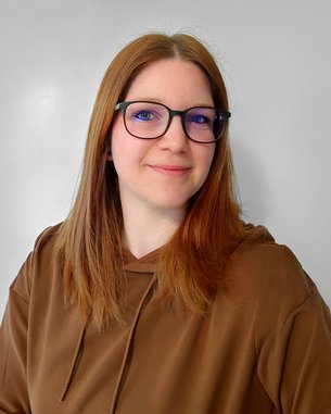 Eine Person mit langen, braunen Haaren und Brille lächelt in die Kamera, trägt einen braunen Hoodie und hat einen neutralen Hintergrund.