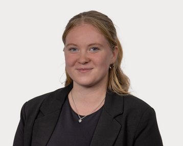 Junge Frau mit blonden Haaren, die in einem schwarzen Blazer lächelt. Der Hintergrund ist neutral und hell.