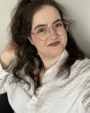 Eine Person mit lockigem, dunklem Haar und Brille lächelt in die Kamera, während sie eine Hand durch ihr Haar fährt. Sie trägt ein weißes Hemd und hat einen neutralen Hintergrund.