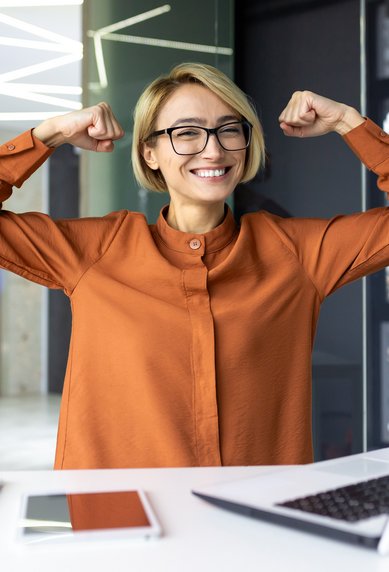 Eine lächelnde Frau mit Brille in einem modernen Büro hebt die Arme in einer Siegerpose. Vor ihr steht ein Laptop auf dem Tisch, daneben liegen ein Notizbuch und Stifte.