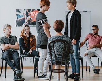 Fernstudium Bachelor Soziale Arbeit Spezialisierung Prekäre Lebenslagen | SRH Fernhochschule Eine Gruppe von Jugendlichen sitzt in einem Raum auf Stühlen. Zwei Jugendliche stehen im Mittelpunkt und scheinen ein Gespräch zu führen, während die anderen aufmerksam zuschauen.