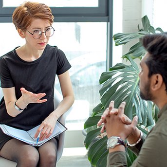 Eine Frau mit Brille sitzt auf einem Stuhl und spricht mit einem Mann, der ihr gegenüber sitzt. Beide scheinen in ein Gespräch vertieft zu sein, umgeben von Pflanzen und natürlichem Licht.