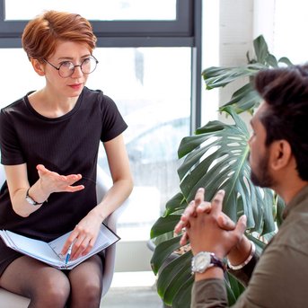 Eine Frau mit Brille sitzt auf einem Stuhl und spricht mit einem Mann, der ihr gegenüber sitzt. Beide scheinen in ein Gespräch vertieft zu sein, umgeben von Pflanzen und natürlichem Licht.