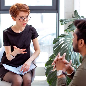 Eine Frau mit Brille sitzt auf einem Stuhl und spricht mit einem Mann, der ihr gegenüber sitzt. Beide scheinen in ein Gespräch vertieft zu sein, umgeben von Pflanzen und natürlichem Licht.