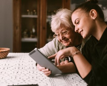 Eine ältere Frau und eine junge Frau sitzen an einem Tisch und schauen gemeinsam auf ein Tablet. Beide lächeln und scheinen sich über den Inhalt des Tablets zu freuen.