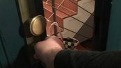 Eine Hand greift nach einem Türknauf, während eine Treppe im Hintergrund sichtbar ist. Ein kariertes Einkaufstasche hängt an der Treppe.
