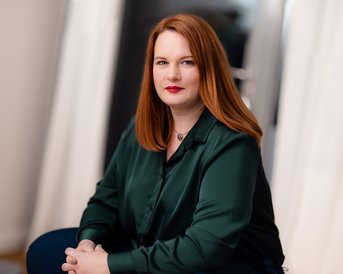 Eine Frau mit langen, roten Haaren sitzt mit verschränkten Armen und einem neutralen Ausdruck. Sie trägt ein grünes Hemd und hat roten Lippenstift auf. Der Hintergrund ist unscharf und hell.