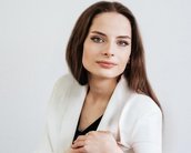 Eine Frau mit langen, braunen Haaren und einem neutralen Ausdruck trägt einen weißen Blazer und posiert vor einem hellen Hintergrund.