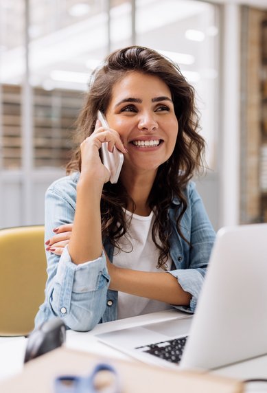 Eine Frau mit lockigem Haar sitzt an einem Schreibtisch, spricht am Telefon und lächelt. Vor ihr steht ein Laptop, umgeben von Kartons und Bürobedarf.