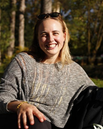 Eine lächelnde Frau mit blonden Haaren trägt einen grauen Pullover und sitzt im Freien, umgeben von Bäumen.