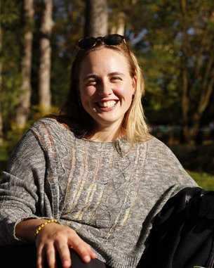 Eine lächelnde Frau mit blonden Haaren trägt einen grauen Pullover und sitzt im Freien, umgeben von Bäumen.