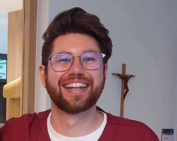 Ein lächelnder Mann in rotem Arztkittel steht neben einer Kaffeemaschine. Er trägt eine Armbanduhr und hat eine Brille auf. Im Hintergrund sind Möbel und eine Wand sichtbar.