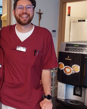 Ein lächelnder Mann in rotem Arztkittel steht neben einer Kaffeemaschine. Er trägt eine Armbanduhr und hat eine Brille auf. Im Hintergrund sind Möbel und eine Wand sichtbar.