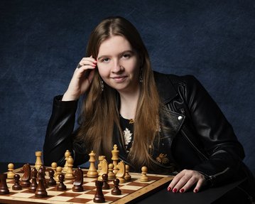 Eine junge Frau mit langen Haaren sitzt an einem Schachbrett, das mit Schachfiguren in verschiedenen Farben besetzt ist. Sie trägt eine schwarze Lederjacke.