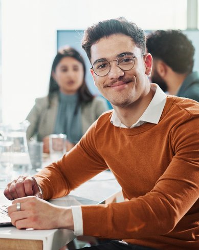 Ein junger Mann mit Brille und braunem Pullover sitzt an einem Tisch mit einem Laptop. Im Hintergrund sind mehrere Personen in einem Besprechungsraum zu sehen.
